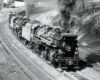 Two double-headed steam locomotives head around a curve hauling a freight train.