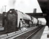 A large steam locomotive pulls a passenger train through a station area.