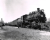 Steam locomotive at the head of a passenger train in a rail yard.