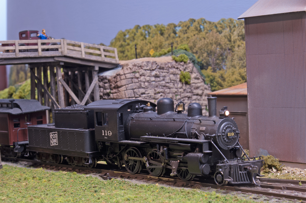 An HO scale 2-6-0 Mogul steam locomotive passes under a wooden road bridge and past a corrugated metal building.