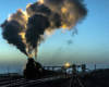 A steam locomotive at the head end of a train billowing smoke and steam in an industrial rail yard.