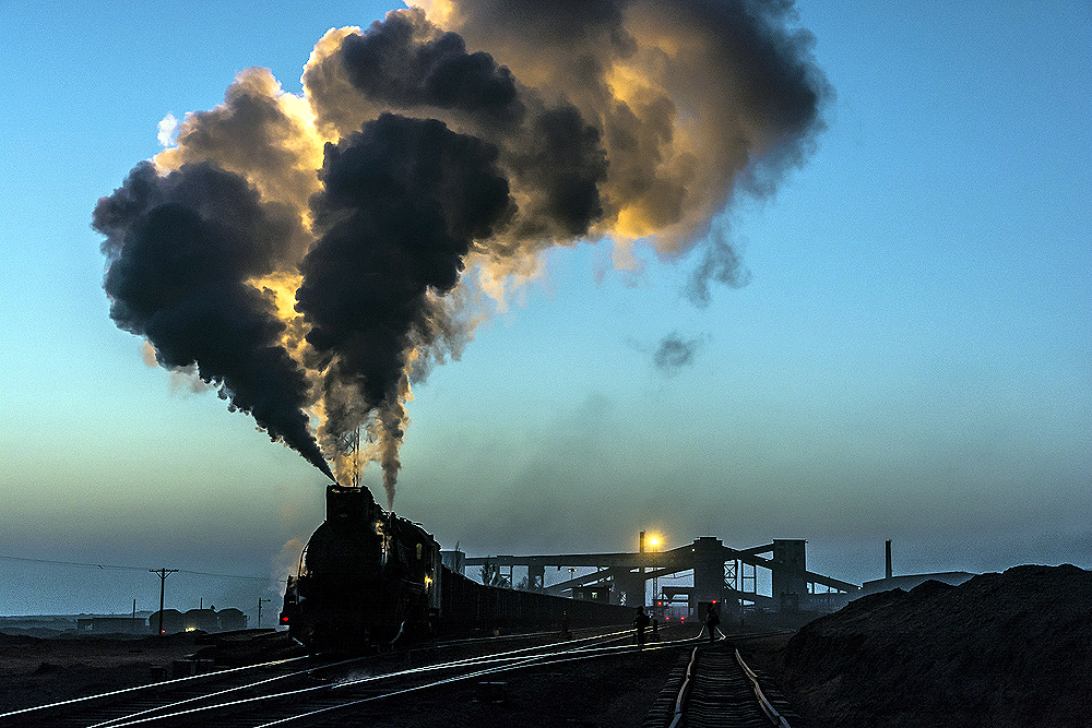 A steam locomotive billows smoke and steam under a twilight sky in an industrial rail yard.