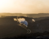 A steam locomotive leads a train through a valley in low-angle sunlight.