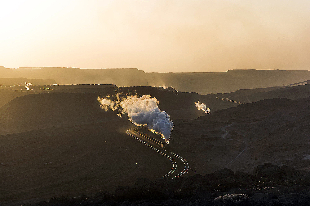 A steam locomotive leads a train through a valley in low-angle sunlight.