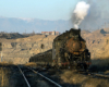 Steam locomotive hauls a train through a mined area. Snow capped mountains in background.