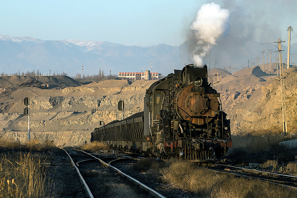 Steam locomotive hauls a train through a mined area. Snow capped mountains in background.
