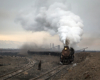 A steam locomotive hauls a train out of a rail yard in an arid desert scene.