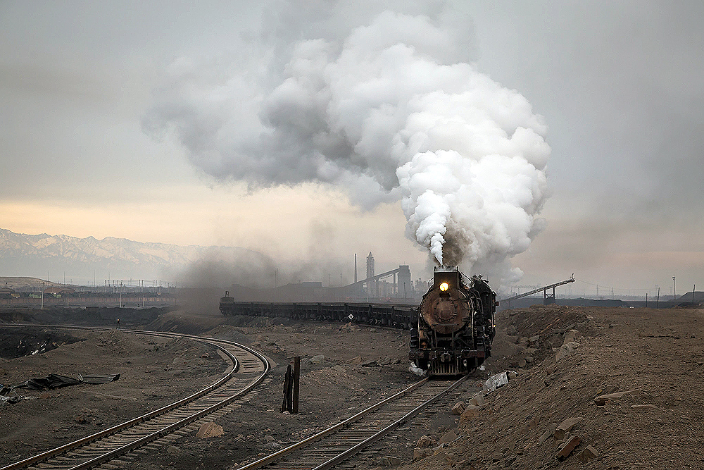 A steam locomotive hauls a train out of a rail yard in an arid desert scene.