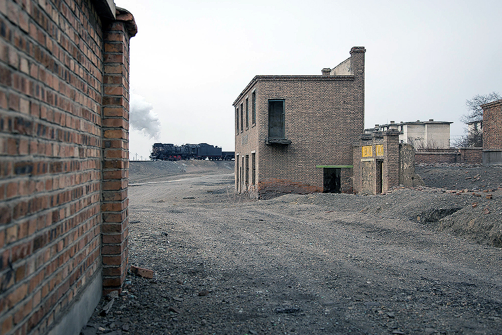 A steam locomotive passes in the background between abandoned buildings.