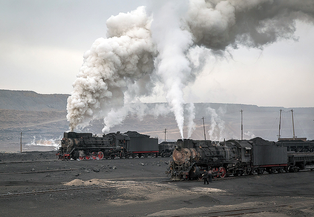 Two steam locomotives paused at the base of an open pit coal mine.