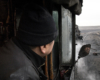 A train crewman peers out his cab window, his face reflected in a small mirror.