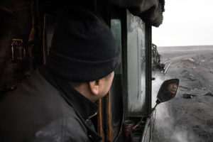 A train crewman peers out his cab window. His face is reflected in a mirror.