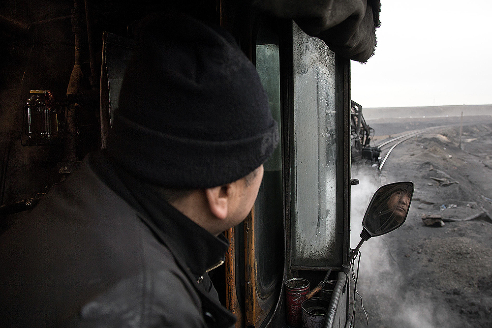 A train crewman peers out his cab window, his face reflected in a small mirror.