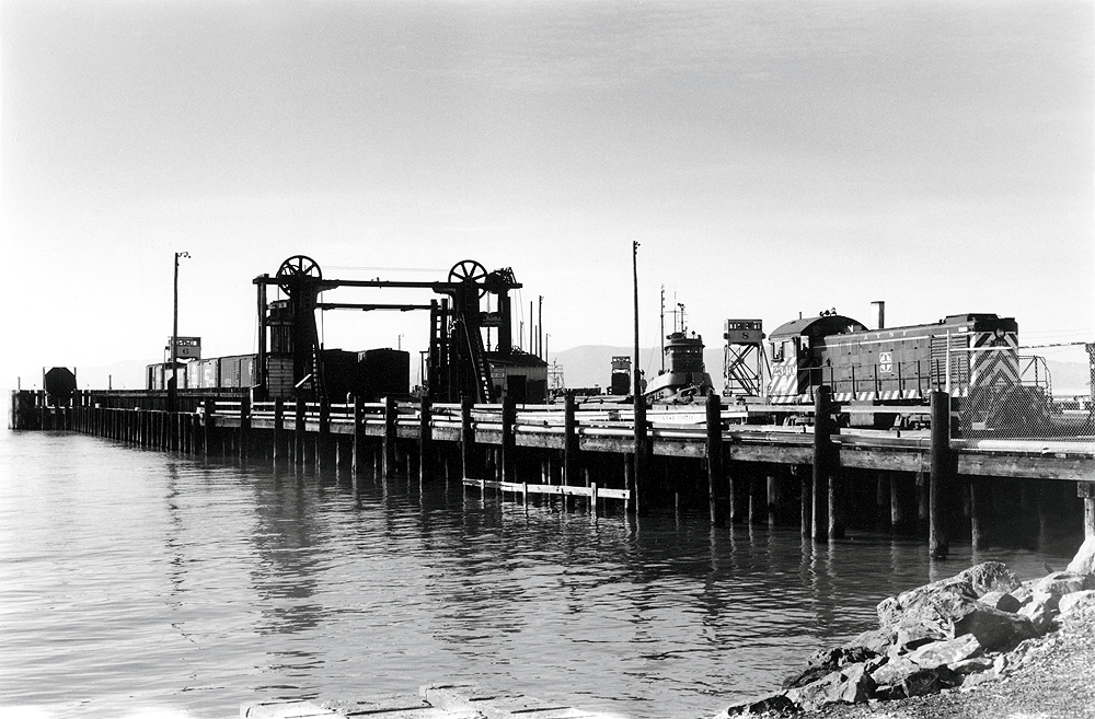 Locomotive works with freight cars on a floating barge connected to land. Black and white.