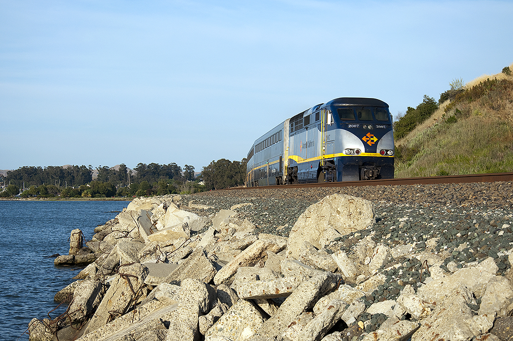 A passenger train on a track on a rocky embankment by a body of water.