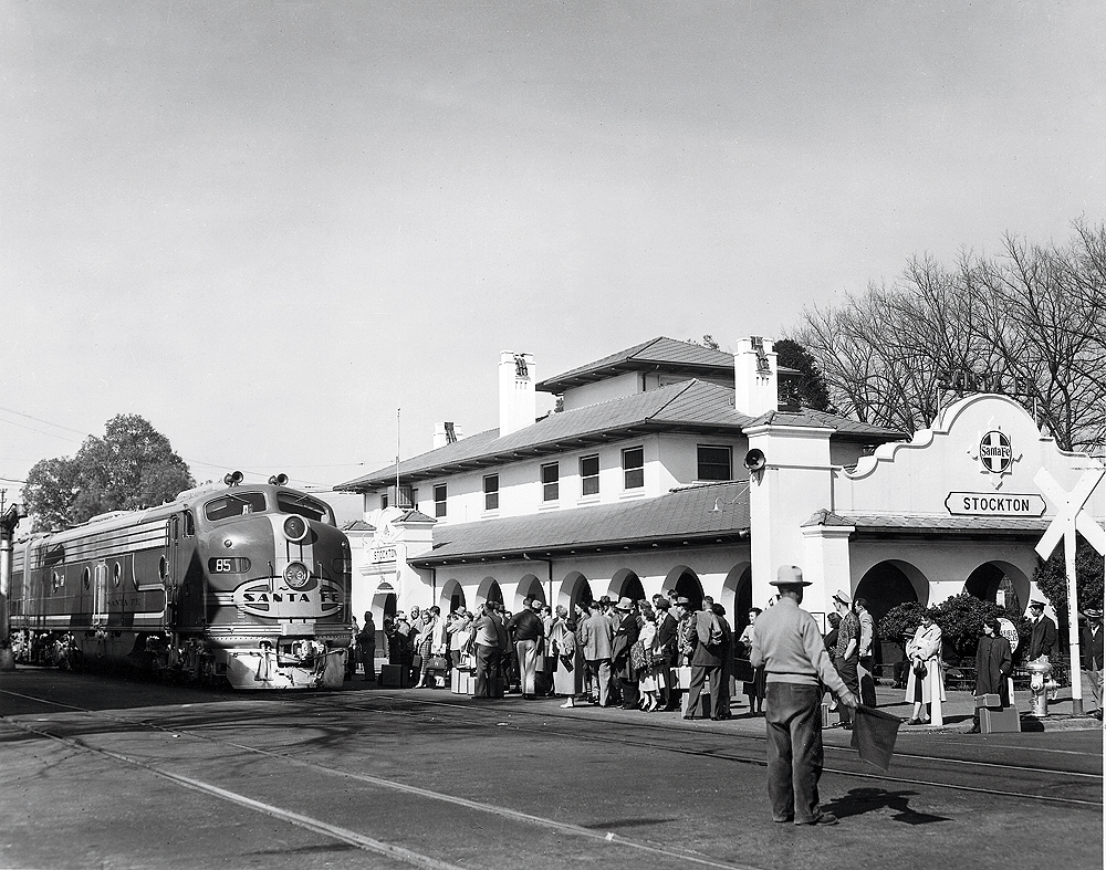 A cab unit-style locomotive in front of a train station surrounded by people. Black and white.