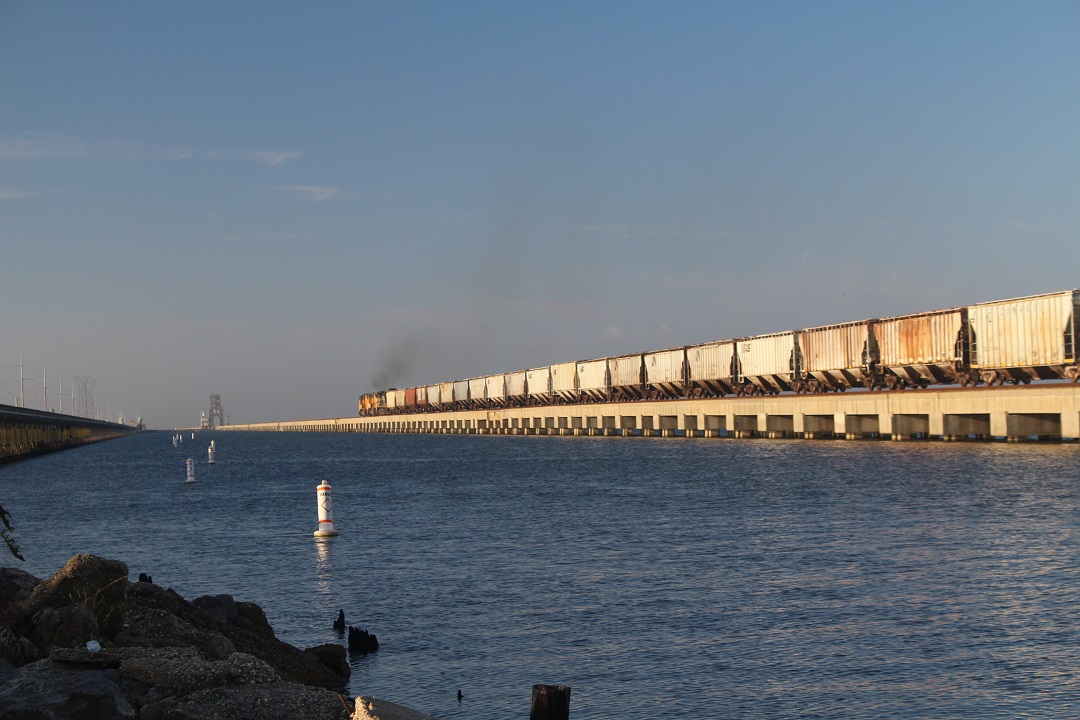 Locomotives hauling a hopper train over a long causeway over a body of water.