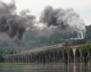 Steam locomotive leading a passenger train over a river on an old stone bridge.