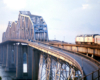 Amtrak diesel locomotives lead a passenger train over a large truss span bridge over a river.