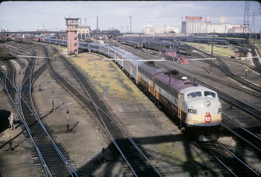 A maroon and gray diesel locomotive leads a passenger train in a rail yard.