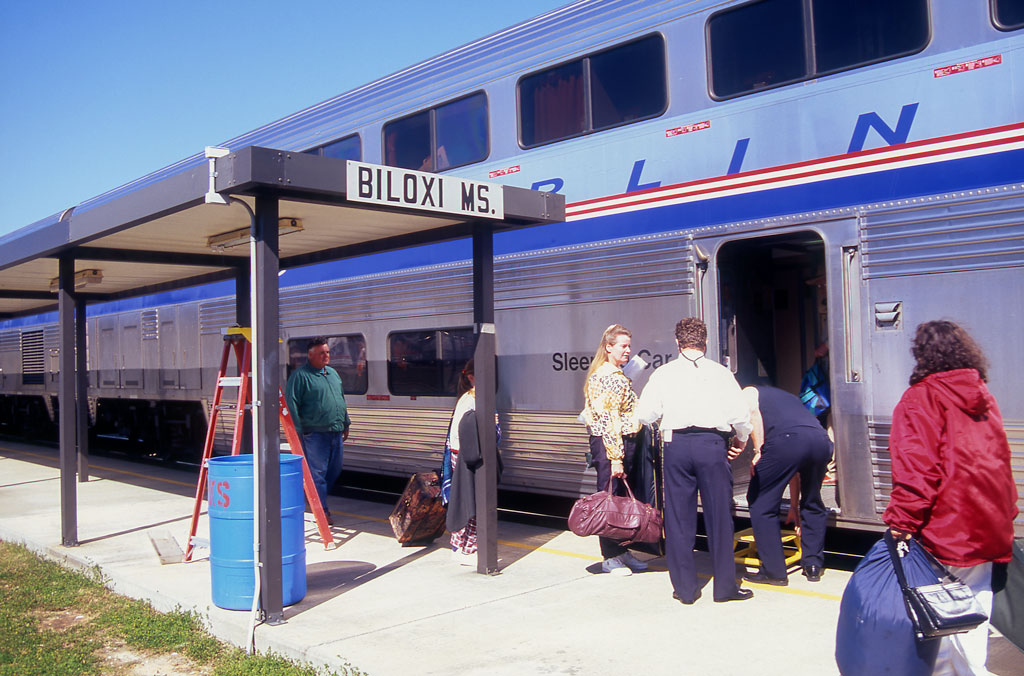 Passengers at a sunny platform board a silver, double-decker passenger car.