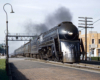 Color image of a bullet shaped steam locomotive at the head of a passenger train.