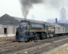 A steel gray steam locomotive at the head of a train in a rail yard.