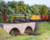 A green Alco diesel leading a freight train rolls onto a stone arch bridge over a creek