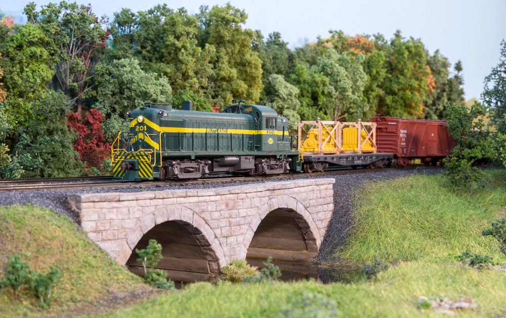 A green Alco diesel leading a freight train rolls onto a stone arch bridge over a creek