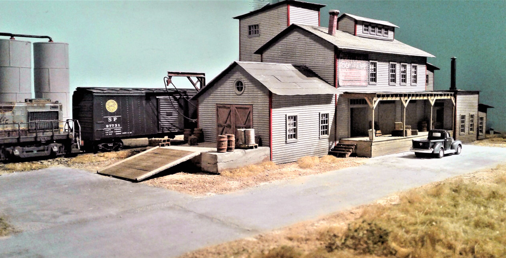 An aged-looking diesel pushes a black steel boxcar to a dock behind a white wood-clapboard factory