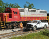 A railroad worker in an orange safety vest stands next to his Hi-Rail truck as he watches a red-and-silver diesel roll by on the other track