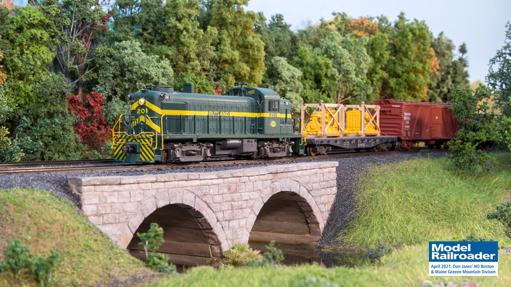 A green diesel locomotive leading a string of freight cars crosses a stone arch bridge over a creek