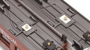 The underside of two HO scale passenger cars, showing bolsters altered with white styrene.