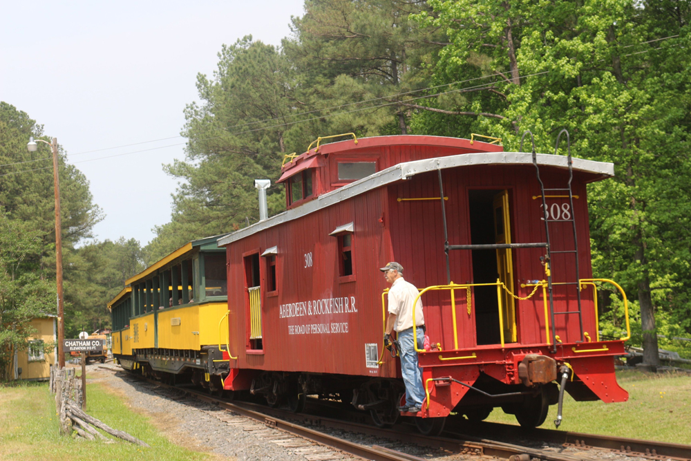 A person rides the steps of a caboose on the back of a train in a green forested landscape.