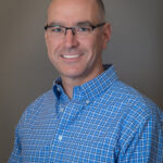 Man with shaved head wearing eyeglasses and blue open-collared shirt.