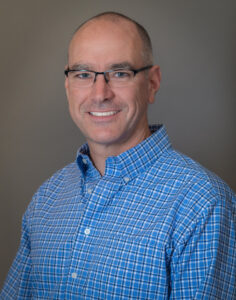 Man with shave head wearing eyeglasses and blue open-collared shirt.