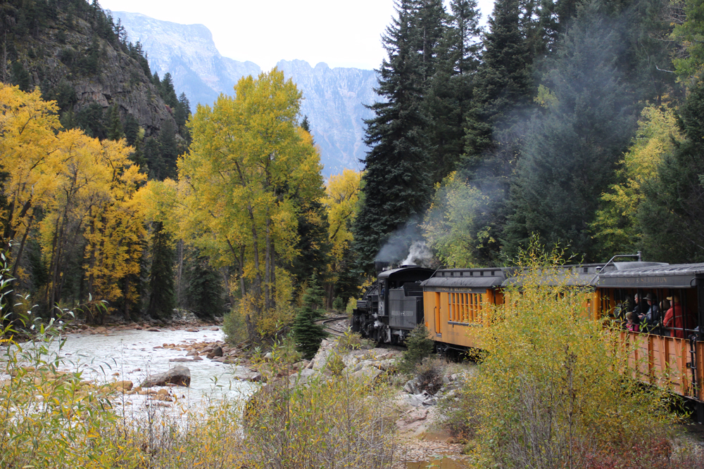 A steam locomotive leads a passenger train along a mountain river valley with trees in autumn colors.