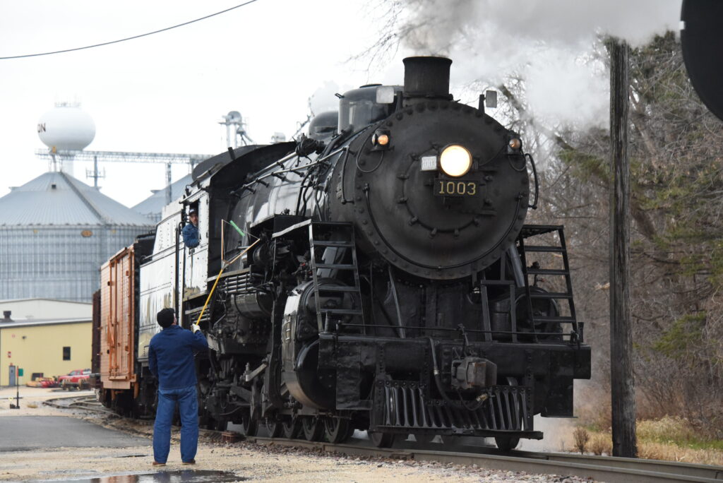 A worker holds a pole next to a steam locomotive moving slowly.