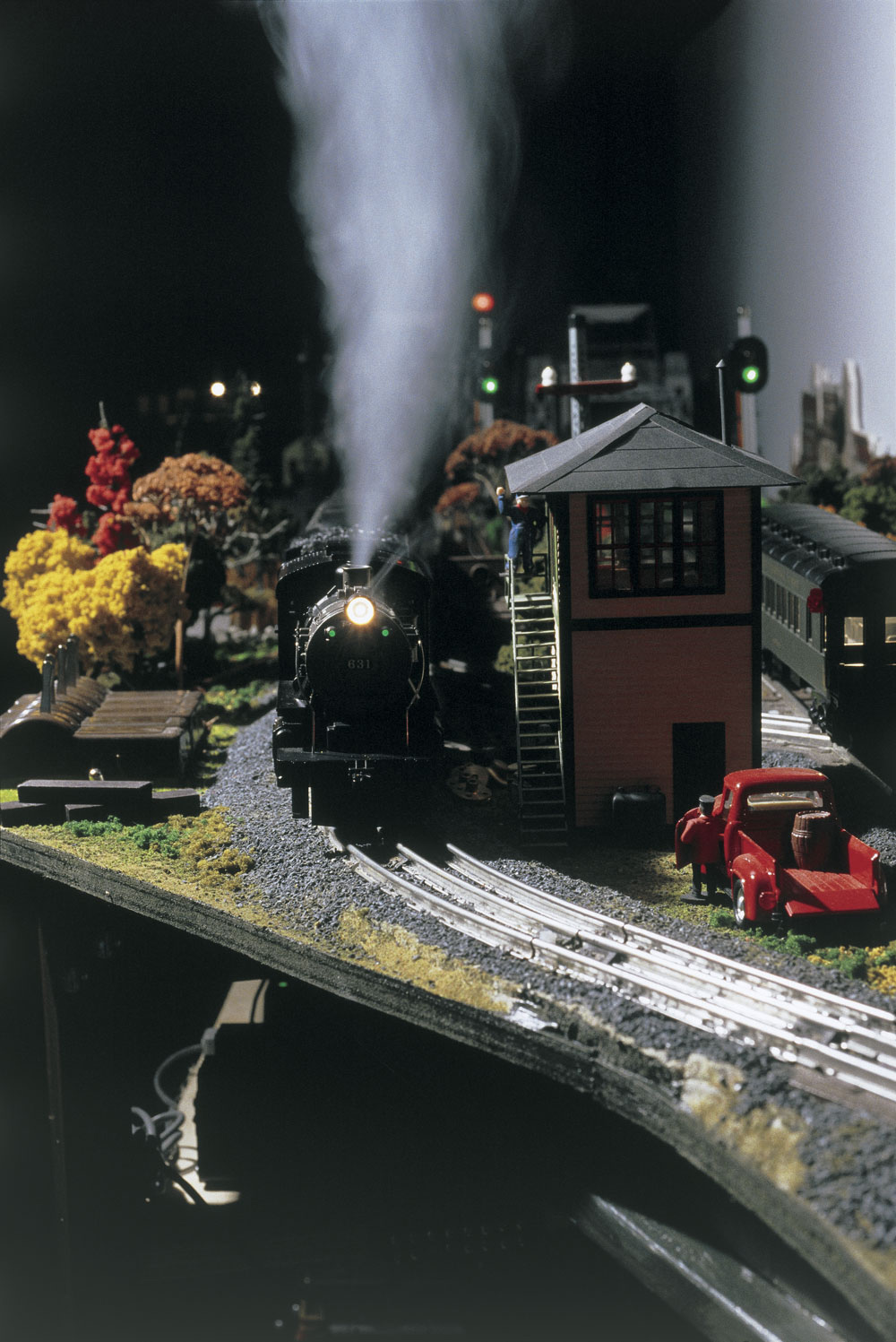 Front of an O gauge model steam locomotive on layout in night operation with smoke coming out of the stack next to interlocking tower.