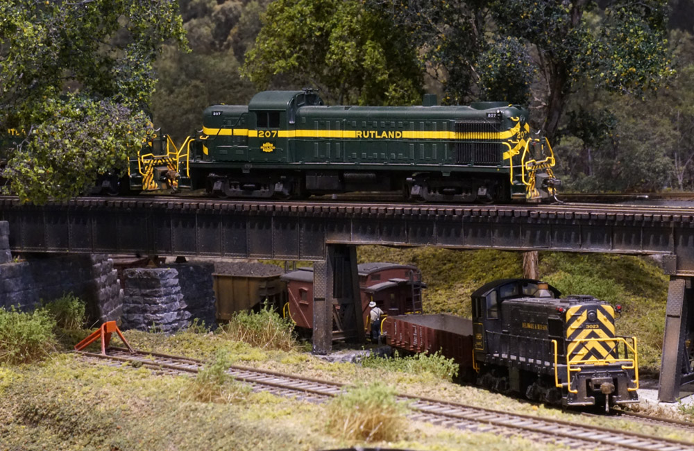 A green road diesel leads a train over a steel girder bridge above a black diesel switcher with its own train