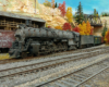 Santa Fe 4-8-4 facing left in front of partial Santa Fe boxcar with route map in front of wood-frame lumber shed, through truss bridge in upper right background