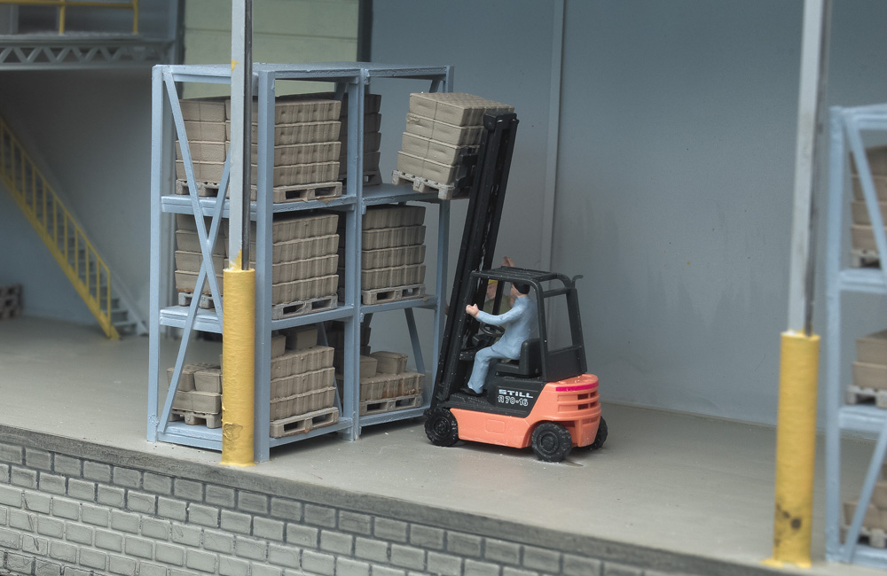 A forklift operator lifts a pallet load of boxes from a high shelf in an HO scale warehouse