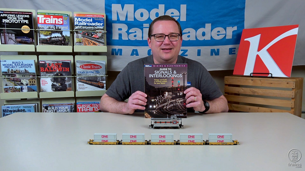 Cody Grivno holds Guide to Signals & Interlockings book, with a silver HO scale caboose with red stripe in front of the book, and a line of N scale Gunderson Maxi-I container cars with containers in front of that