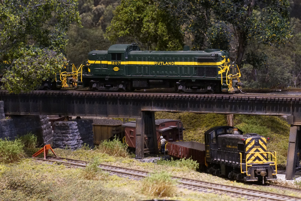 A green road diesel leads a train over a steel girder bridge above a black diesel switcher with its own train