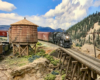 A steam locomotive approaches a trestle as another fills up at the water tank below