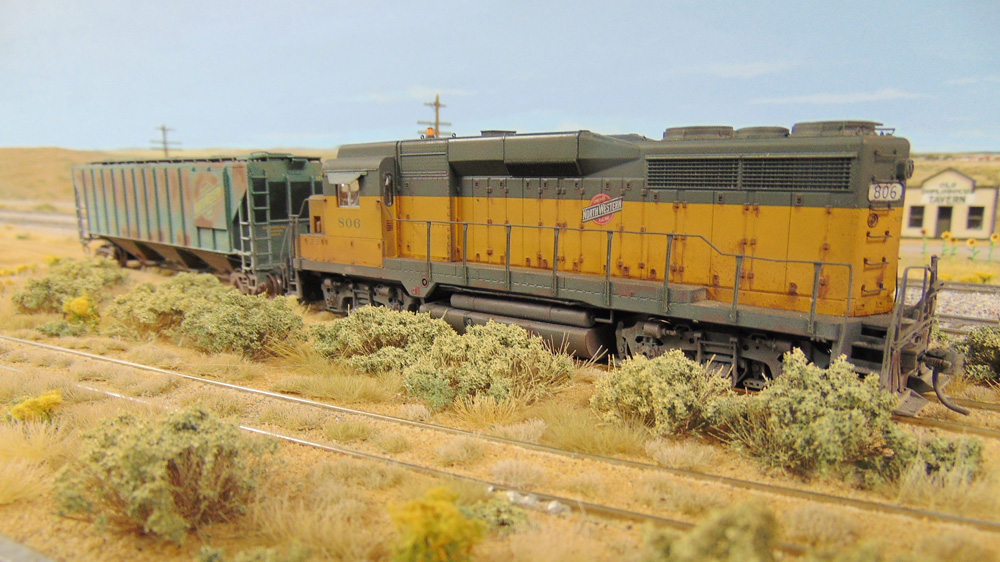 : A rusty yellow-and-green diesel and a worn green covered hopper roll down an equally neglected spur track on the prairie