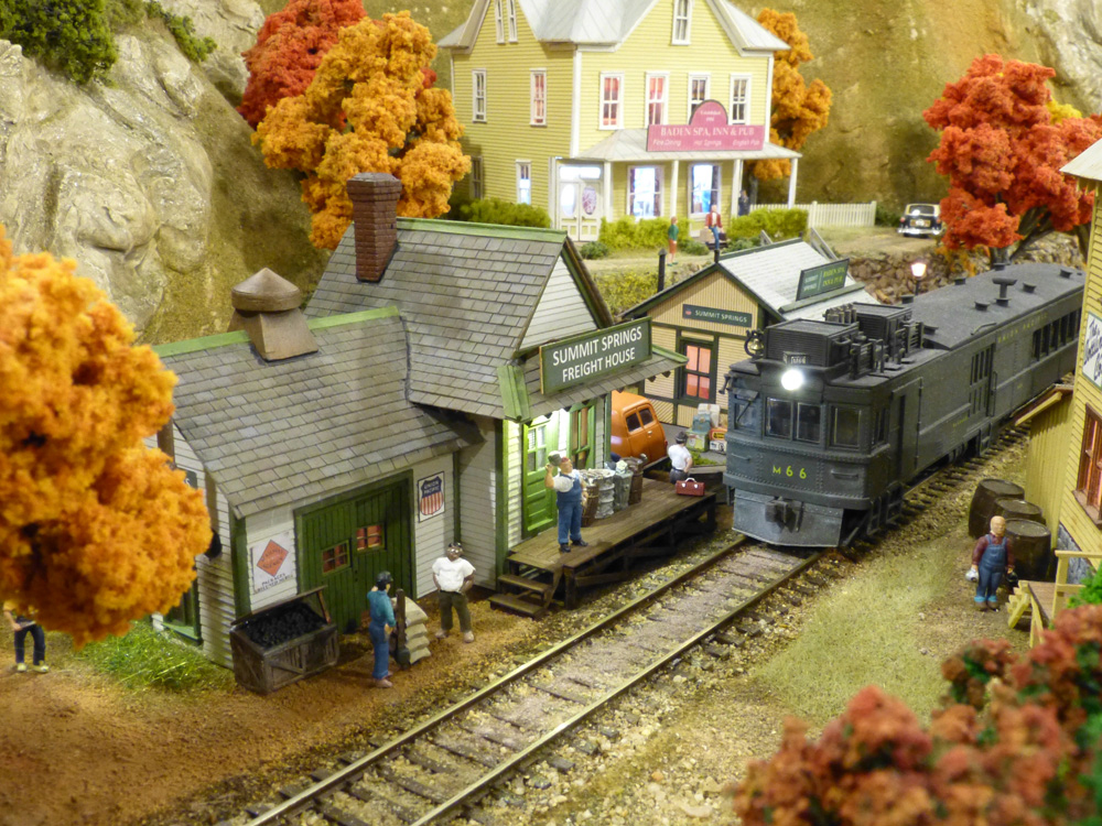 A green “Doodlebug” motorcar pulls up to the platform at a small, yellow, wood-frame depot