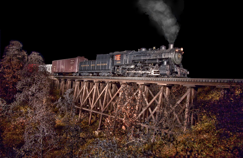 A steam locomotive is dramatically lit from the side as it crosses a wood trestle at night