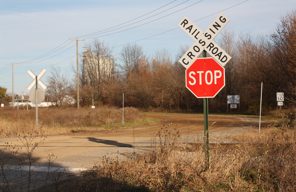 A crossbuck with a stop sign stands at a rural grade crossing with a grain elevator in the distance