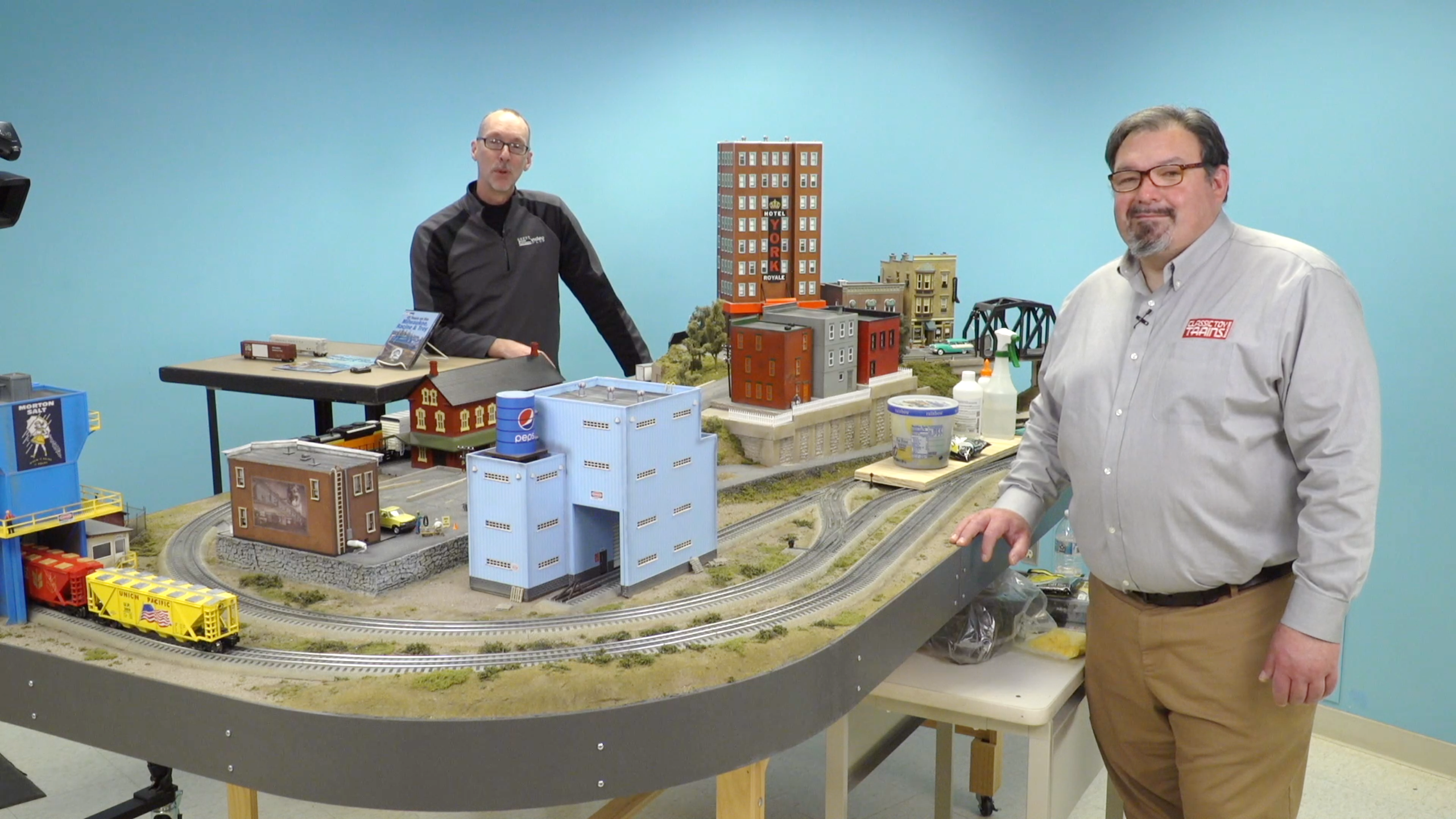 Two men standing on either side of a table-based toy train layout.